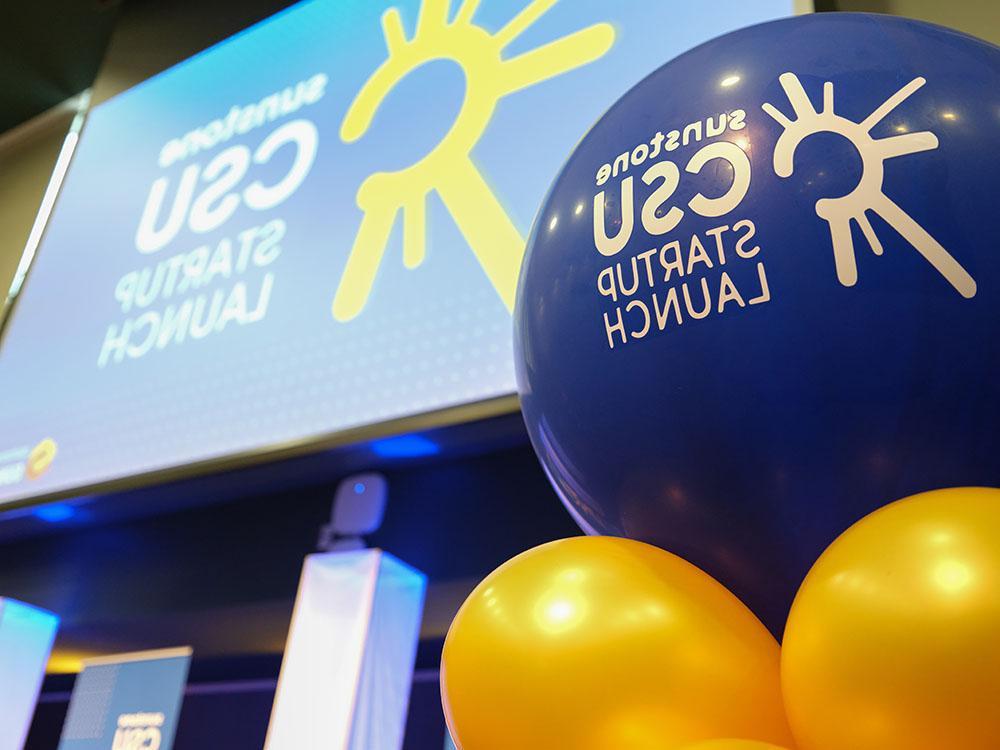 A blue balloon with a starburst logo and the words "Sunstone CSU Startup Competition" in the foreground, with a screen on which is projected the same logo on a blue background behind it.