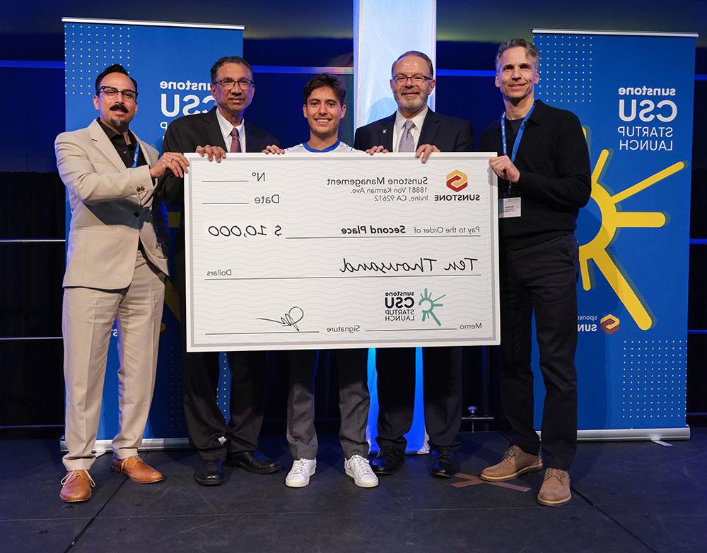 A male, college-age startup founder poses onstage with two sponsors, two hosts, and a giant check for $10,000