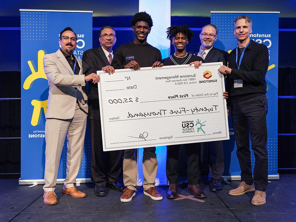 Two male college-age startup founders pose onstage with two sponsors, two hosts, and a giant check for $25,000