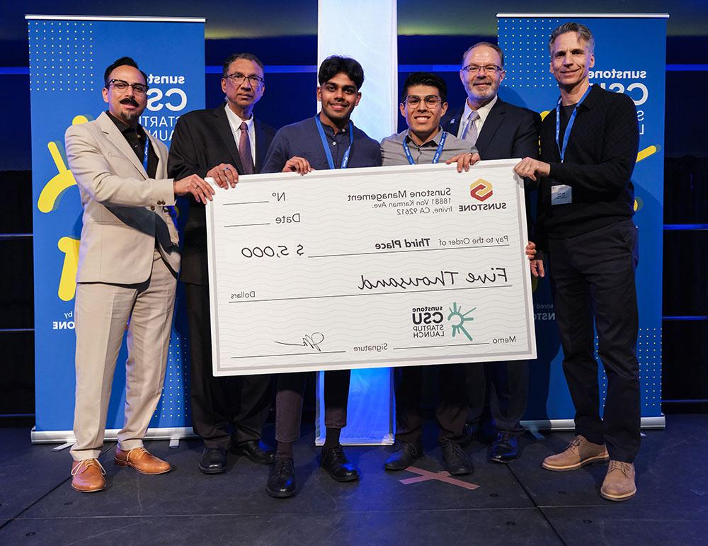 Two college-age startup founders pose onstage with two sponsors, two hosts, and a giant check for $5,000