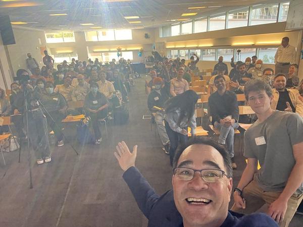 A dark haired man dressed in a sport coat and tshirt, laughing, takes a selfie with an amber-toned roomful of people behind him. 